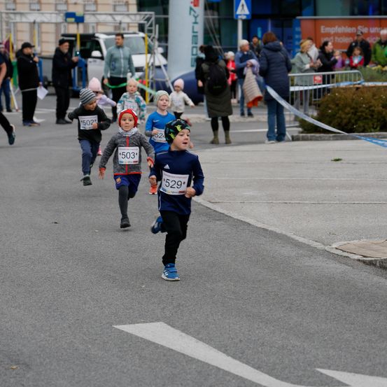 Mehrere Kinder laufen in einem Rennen. Sie tragen nummerierte Trikots. Zuschauer beobachten sie von den Seiten. Die Straße hat weiße Markierungen und Absperrungen.