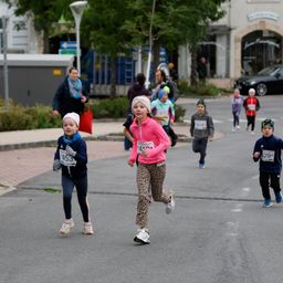 Eine Gruppe von Kindern mit nummerierten Trikots läuft auf einer Straße, mit Zuschauern und Straßenlaternen in der Nähe.
