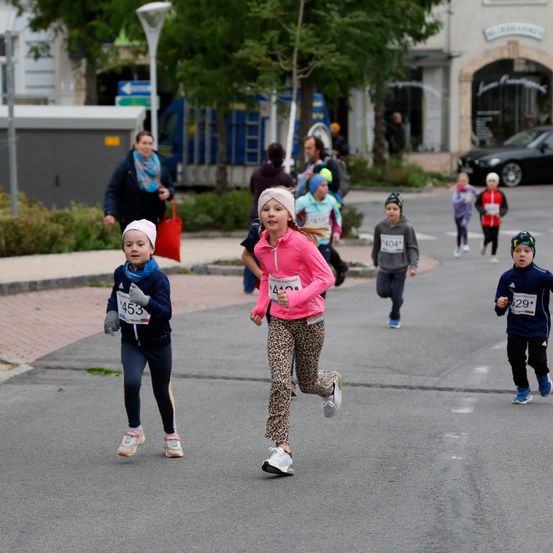 Eine Gruppe von Kindern mit nummerierten Trikots läuft auf einer Straße, mit Zuschauern und Straßenlaternen in der Nähe.