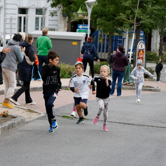 Eine Gruppe junger Kinder läuft auf einer Stadtstraße, während mehrere Menschen stehen und herumlaufen, einige von ihnen klatschen.