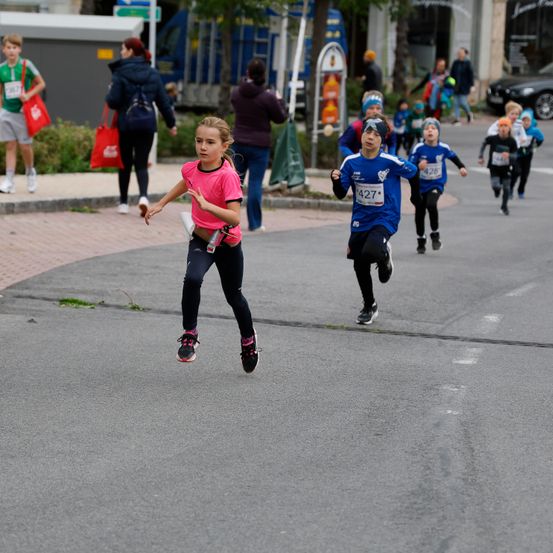 Mehrere Kinder laufen bei einem Rennen auf einer Straße, angeführt von einem jungen Mädchen in einem rosa Shirt. Zuschauer und Läufer befinden sich auf den Gehwegen, und im Hintergrund sind Gebäude mit Geschäften zu sehen.