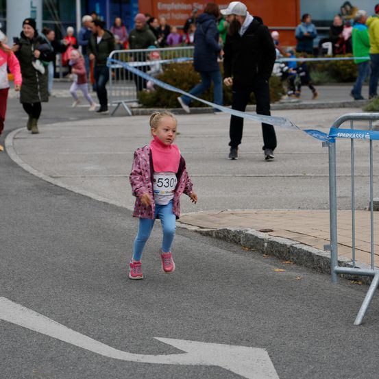 Ein kleines Mädchen in einer pinkfarbenen Jacke mit der Nummer 530 läuft auf der Straße. Hinter ihr steht ein Mann in einer schwarzen Jacke bei einer Barriere mit blauem Band. Mehrere Menschen gehen auf dem Bürgersteig.