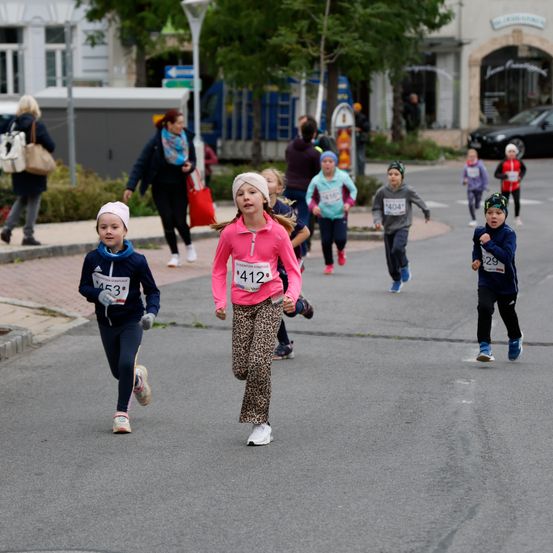 Kinder nehmen an einem Laufwettbewerb auf einer asphaltierten Straße teil, mit Geschäften an der Seite. Das Mädchen in Pink führt, während andere dicht folgen. Zuschauer stehen auf dem Bürgersteig.