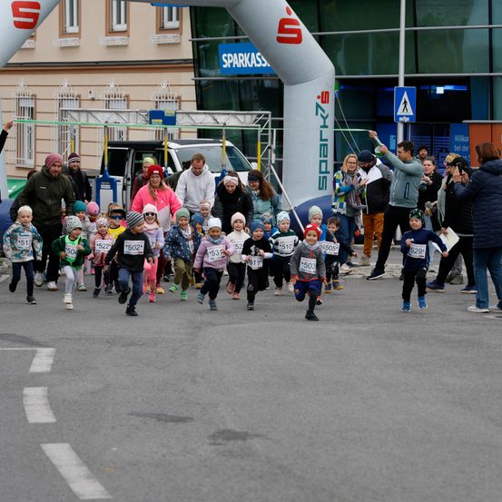 Eine Gruppe von Kindern läuft in einem Rennen und trägt Nummern auf ihren Trikots. Zuschauer feuern sie an. Hinter ihnen steht ein weißer Bogen mit dem Wort Sparkas. Im Hintergrund sind Gebäude und geparkte Autos zu sehen.
