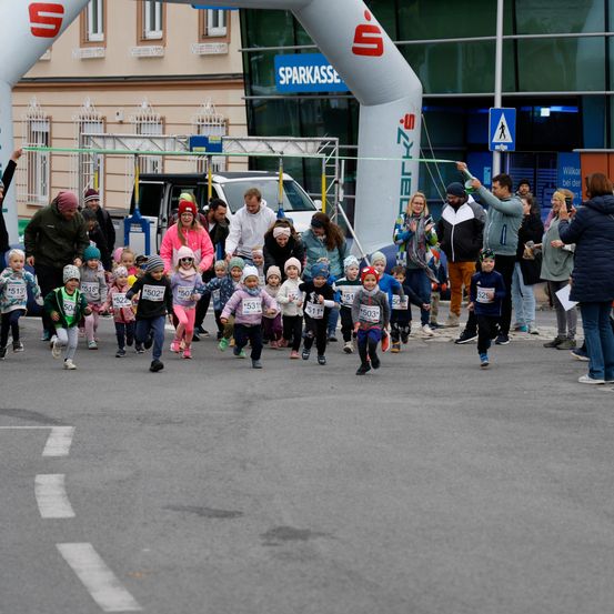 Eine Gruppe von Kindern mit Nummern läuft durch einen Bogen bei einem Rennen. Zuschauer beobachten von hinten.