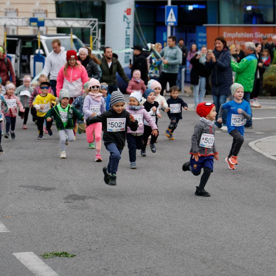 Kinder, die an einem Rennen teilnehmen, mit Nummern auf ihren Trikots, laufen auf einer asphaltierten Straße mit weißen Markierungen. Dahinter stehen Erwachsene, einige halten Kameras und machen Fotos.