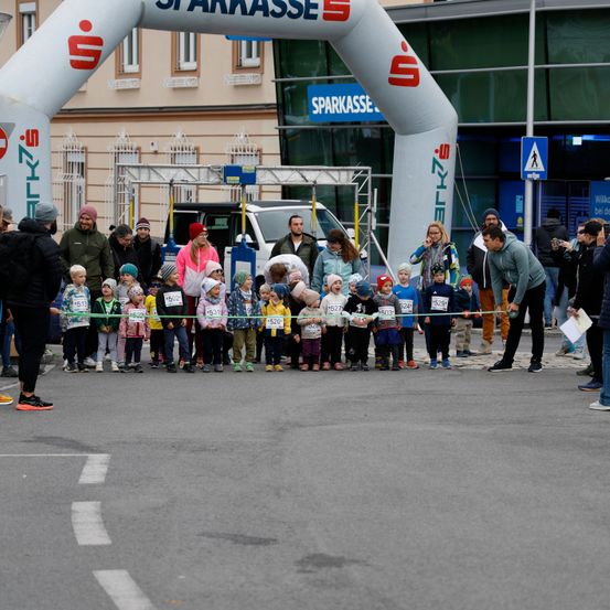 Kinder in Winterkleidung stehen hinter einem grünen Band beim Start eines Marathons. Ein Mann hält das Band mit einer Frau und mehreren Leuten im Hintergrund.