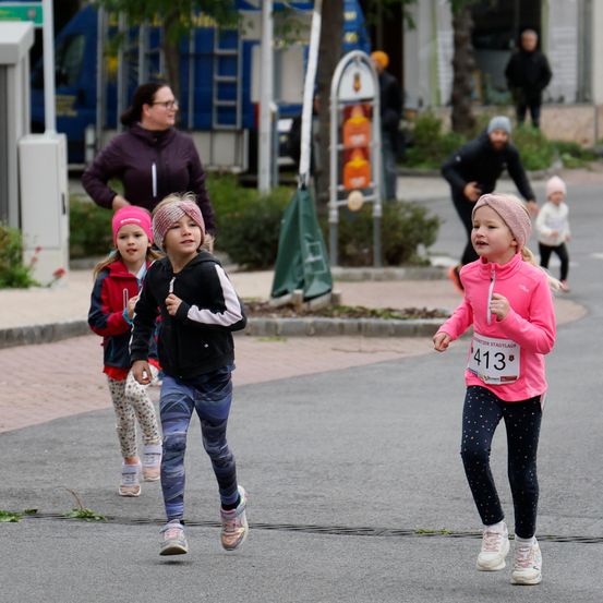 Drei Kinder laufen in einem Rennen auf einer Straße. Ein Mädchen in Pink führt, gefolgt von zwei anderen. Eine erwachsene Frau ist hinter ihnen. Im Hintergrund gehen Leute auf dem Bürgersteig.