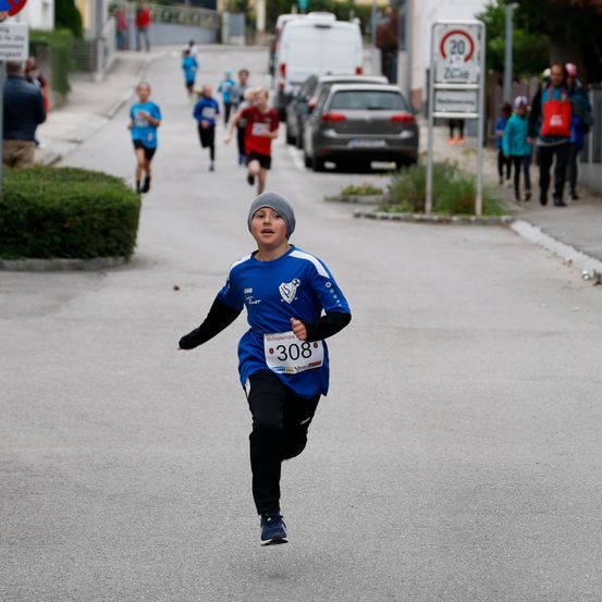 Ein Junge mit der Nummer 308 läuft bei einem Rennen. Mehrere Menschen laufen hinter ihm. Es gibt geparkte Autos und Menschen am Straßenrand.