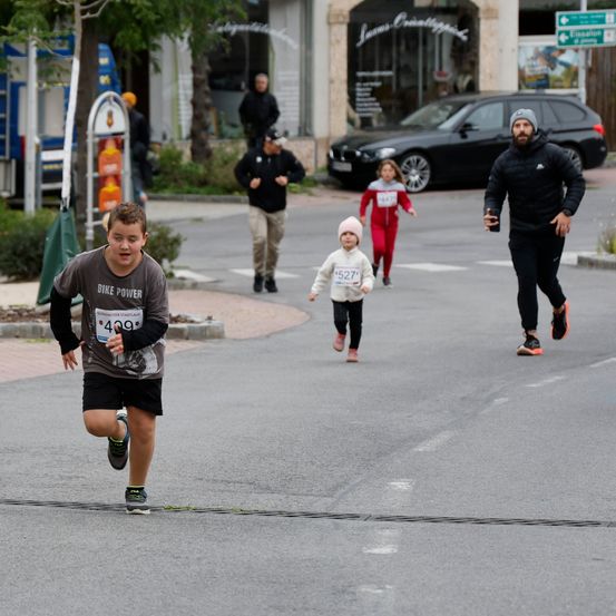 Ein Junge in einem grauen Shirt läuft in einem Marathon. Hinter ihm joggt ein Mann in schwarzer Kleidung. Auf der linken Seite läuft ein kleines Mädchen in pinker Kleidung. Ein schwarzes Auto ist am Straßenrand geparkt.