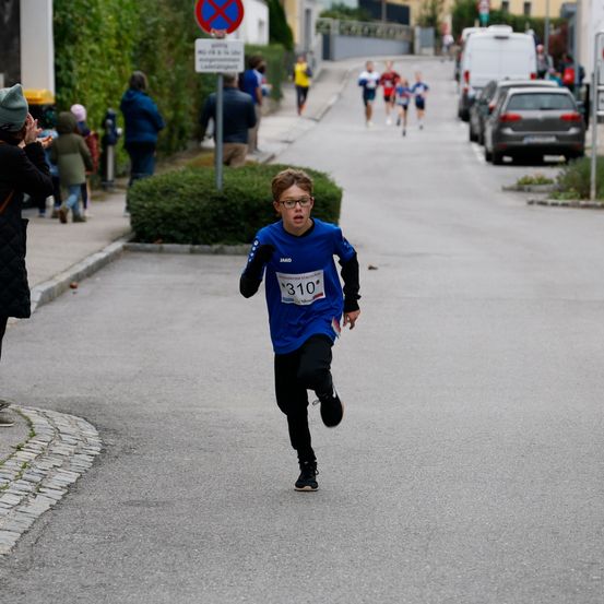 Ein junger Junge läuft bei einem Rennen auf der Straße, trägt eine Brille und ein blaues Trikot mit der Nummer 310. Leute beobachten ihn vom Bürgersteig aus. Im Hintergrund folgen weitere Läufer, und Autos sind am Straßenrand geparkt.