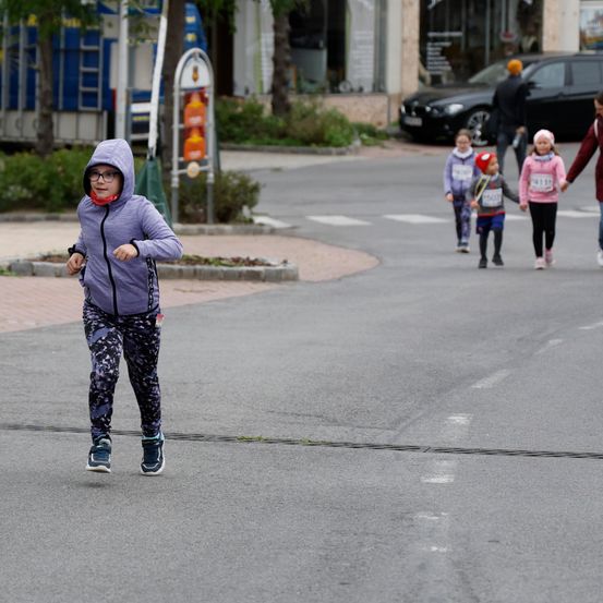 Ein junges Mädchen läuft mit einer Brille und einer lila Kapuze über eine Straße mit einem Zebrastreifen. Dahinter läuft eine Gruppe von Kindern mit Nummern auf ihren Trikots. Ein schwarzer Wagen ist geparkt, und eine Person geht auf dem Bürgersteig.