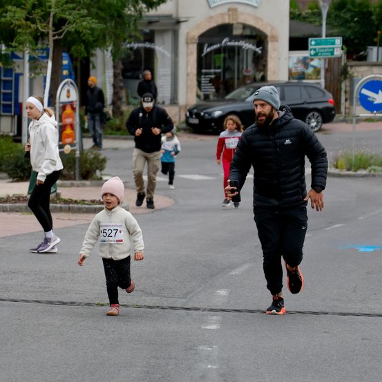 Mehrere Menschen laufen auf einer Straße. Ein junges Mädchen in einem weißen Shirt mit der Nummer 527 läuft. Ein Mann läuft vor ihnen. Ein weiterer Mann läuft hinter ihnen. Ein Mädchen geht auf dem Bürgersteig.