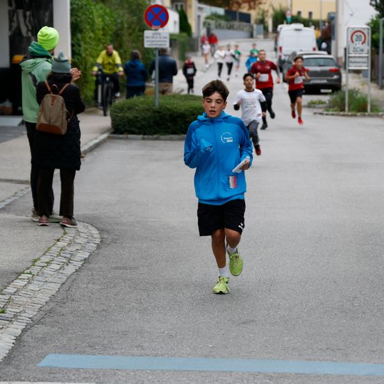 Ein junger Junge in einer blauen Jacke und schwarzen Shorts läuft in einem Rennen mit anderen Kindern und Erwachsenen an der Seite.