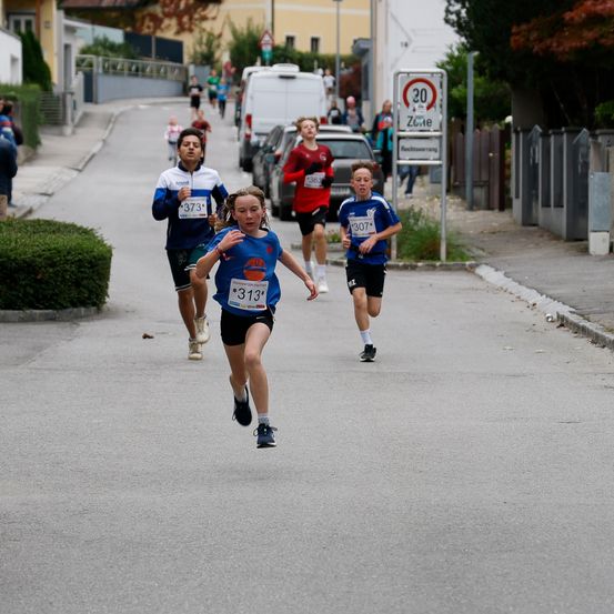 Drei Läufer nehmen an einem Rennen auf einer Straße teil. Die Läuferin in Führung hat die Nummer 313. Dahinter folgen zwei weitere Läufer mit den Nummern 373 und 307. Die Straße hat geparkte Autos und ein Schild mit der Geschwindigkeitsbegrenzung von 20 km/h.