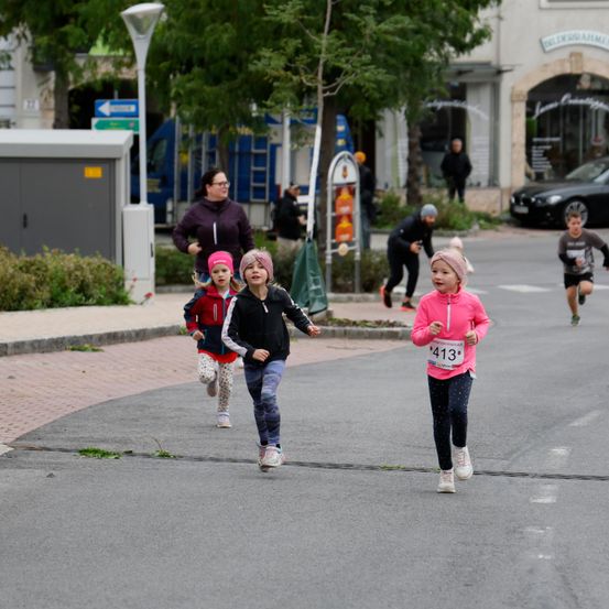 Mehrere Kinder laufen auf einer Straße und tragen nummerierte Startnummern. Eine Frau steht in der Nähe und beobachtet sie. Bäume und Geschäfte sind im Hintergrund zu sehen.