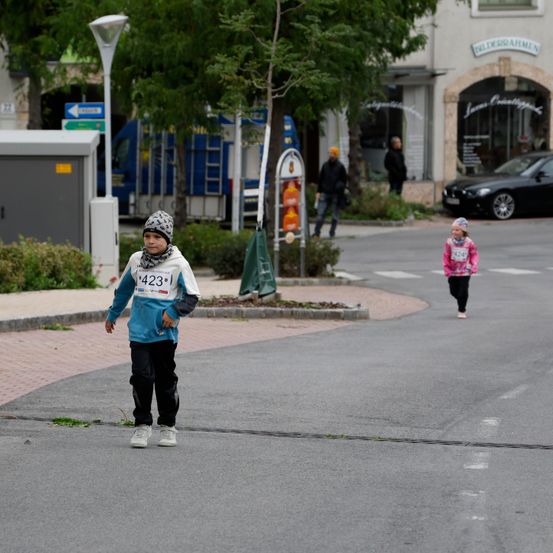 Ein Junge mit der Nummer 423 läuft auf der Straße, während ein Mädchen in einem rosa Shirt folgt. Im Hintergrund befinden sich Gebäude und geparkte Autos.