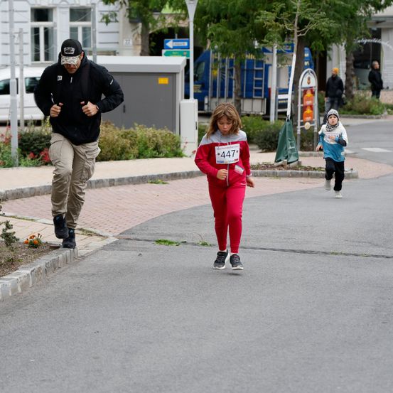 Ein Mann und ein Mädchen in Rot laufen auf der Straße. Der Mann trägt eine schwarze Jacke und Hose, und das Mädchen hat die Nummer 447 auf ihrem Shirt.