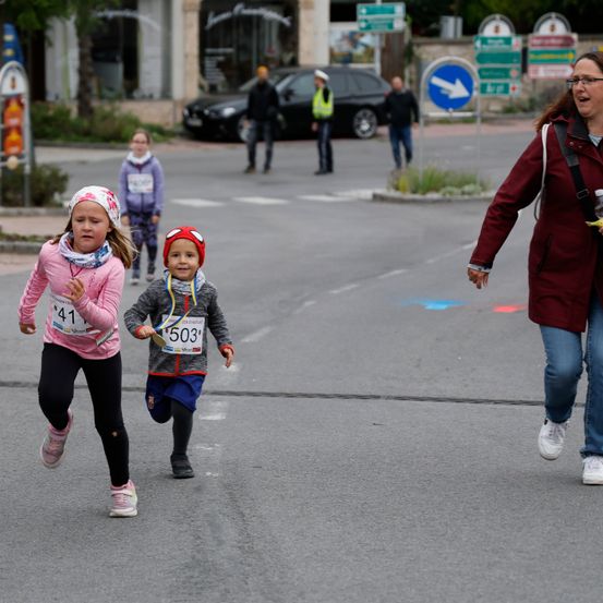 Kinder laufen in einem Rennen. Ein Mädchen mit der Nummer 41 und ein Junge mit der Nummer 503 laufen auf der Straße. Eine Frau und ein Mädchen laufen im Hintergrund.