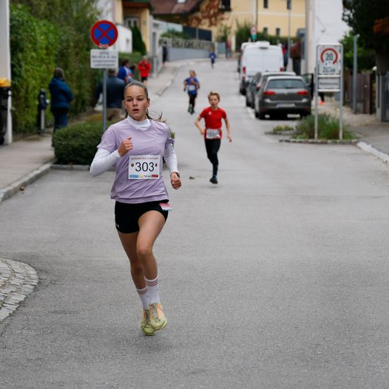 Ein Mädchen mit der Nummer 303 auf dem Trikot läuft bei einem Rennen auf der Straße, mit einem anderen Mädchen in einem roten Trikot hinter ihr. Im Hintergrund sind geparkte Autos und ein Gebäude.