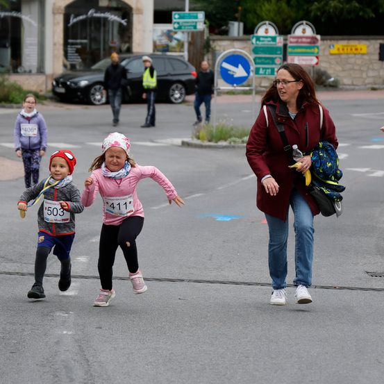 Kinder laufen in einem Rennen mit Nummern auf ihren Trikots. Eine Frau beobachtet in der Nähe. Im Hintergrund befinden sich Gebäude und geparkte Autos.