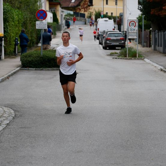 Ein junger Mann läuft in einem Rennen. Er trägt ein weißes T-Shirt mit der Nummer 374. Hinter ihm laufen mehrere Personen. Am Straßenrand sind Autos geparkt.