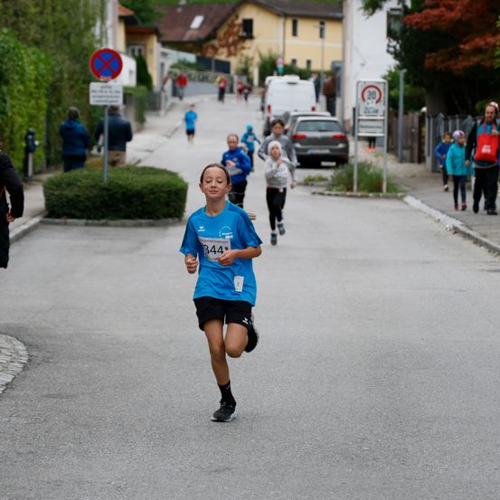 Ein junger Läufer sprintet während eines Rennens eine Straße hinunter, gefolgt von anderen Läufern. Auf dem Bürgersteig befinden sich Zuschauer.