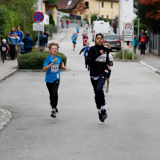 Kinder nehmen an einem Rennen auf einer Straße teil, ein Läufer im blauen Shirt trägt die Startnummer 341 und ein anderer im schwarzen Shirt die Startnummer 349. Zuschauer beobachten von den Seiten.
