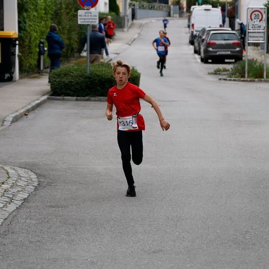 Ein Junge mit der Nummer 315 läuft in einem Rennen die Straße hinunter, mit geparkten Autos und Fußgängern auf dem Bürgersteig.