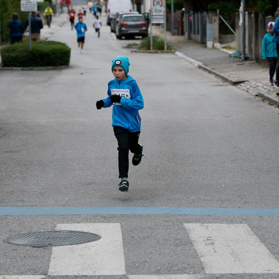 Ein junger Junge in einer blauen Jacke und Mütze läuft bei einem Marathon auf einer Straße. Mehrere Läufer folgen ihm.