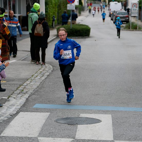 Ein junger Läufer in einem blauen Trikot mit der Nummer 384 sprintet während eines Rennens die Straße hinunter. Zuschauer beobachten es am Bürgersteig.
