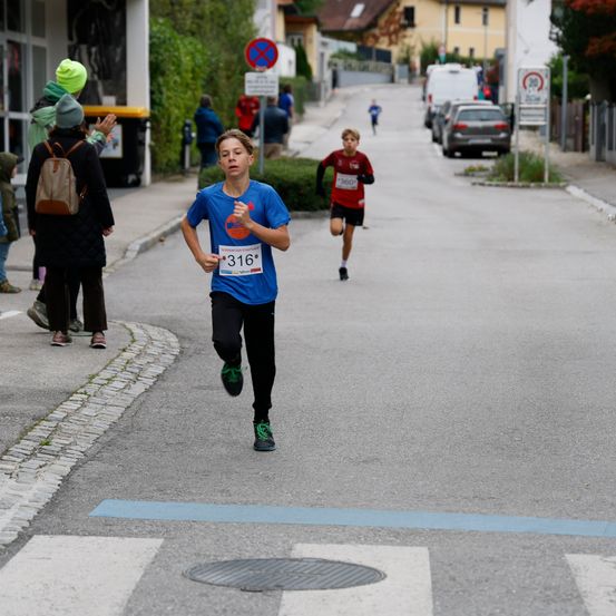 Ein Junge mit der Nummer 316 läuft in einem Rennen die Straße hinunter. Eine Person mit der Nummer 360 läuft hinter ihm. Leute gehen auf dem Bürgersteig.