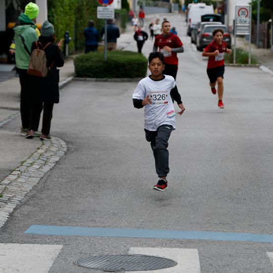 Ein junger Junge mit der Nummer 326 läuft in einem Rennen mit anderen auf einer Straße. Zuschauer beobachten vom Bürgersteig.