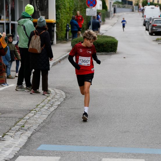 Ein Junge in einem roten Shirt mit der Nummer 360 läuft auf der Straße, während mehrere Menschen vom Bürgersteig aus zuschauen.