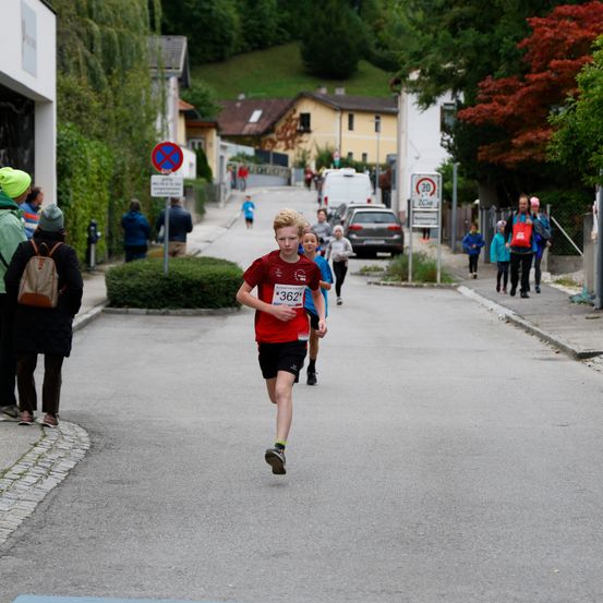 Ein Junge läuft auf der Straße einen Marathon. Mehrere Leute beobachten ihn. Einige Autos sind geparkt. Es gibt Bäume, Häuser und Büsche.