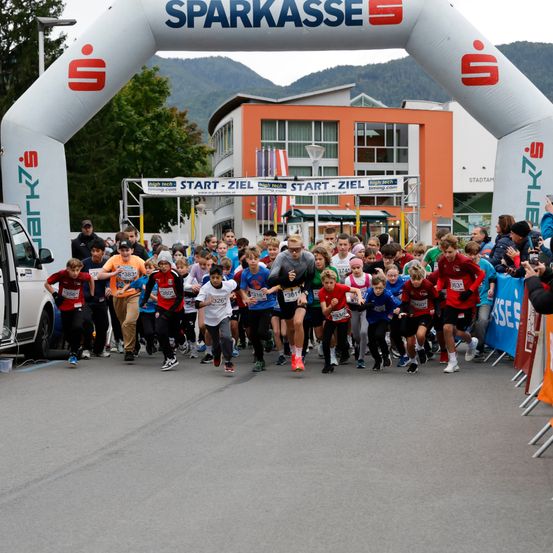 Eine Gruppe von Kindern nimmt an einem Marathon unter einem weißen Torbogen mit 'SPARKASSE' teil. Zuschauer beobachten von den Seitenlinien, und Gebäude sind im Hintergrund sichtbar.