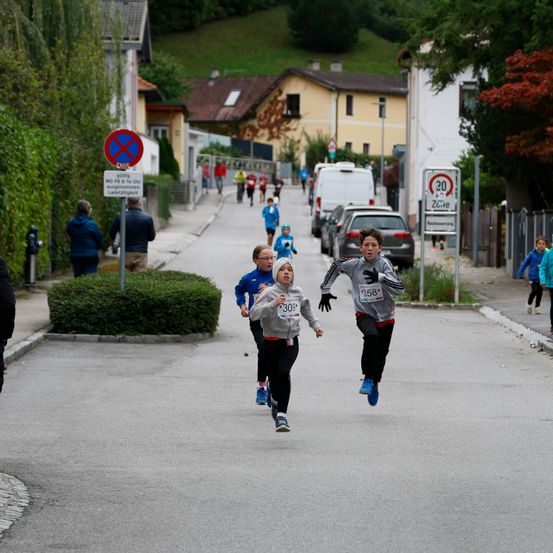 Eine Gruppe von Kindern und Erwachsenen nimmt an einem Laufevent teil. Ein Geschwindigkeitsbegrenzungsschild von 30 ist sichtbar. Die Teilnehmer tragen nummerierte Shirts. Gebäude und geparkte Autos säumen die Straße.