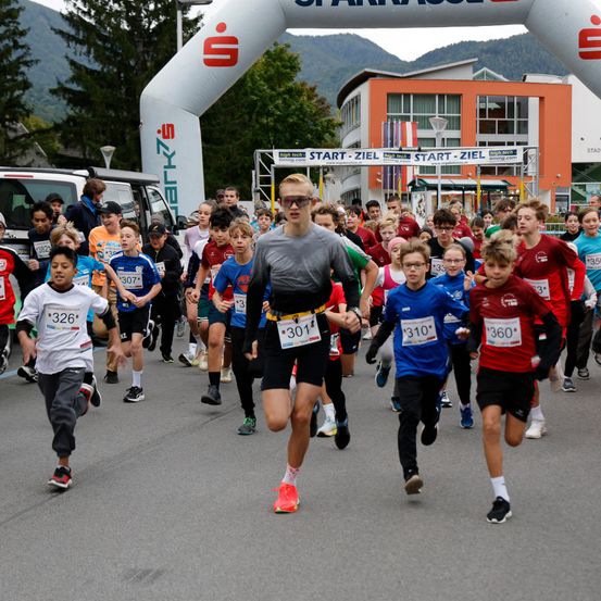 Läufer unterschiedlichen Alters nehmen an einem Marathon teil und passieren ein Tor mit Event-Branding. Eine Menge Zuschauer beobachtet.