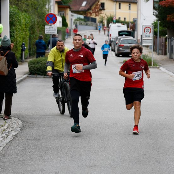 Läufer nehmen an einem Rennen teil, einer trägt die Nummer 3, ein anderer die Nummer 365, ein Radfahrer folgt. Zuschauer beobachten von der Seite.