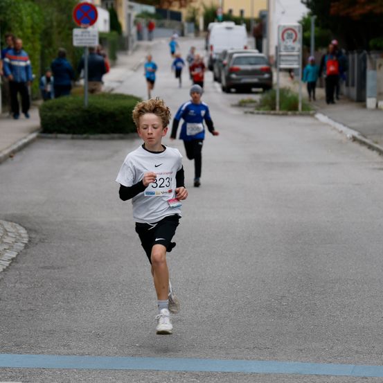 Ein Junge läuft in einem Rennen mit der Nummer 323 auf seinem Shirt, während andere hinter ihm auf einer Straße mit geparkten Autos und zuschauenden Menschen folgen.