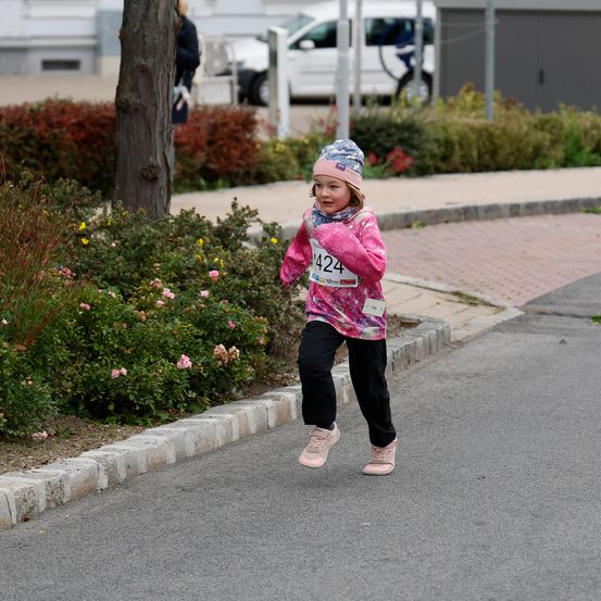 Ein junges Mädchen läuft auf der Straße, Nummer 424 auf ihrem Shirt. Die Straße ist von Grünpflanzen und einem Baum gesäumt. Im Hintergrund ist ein weißer Wagen geparkt.