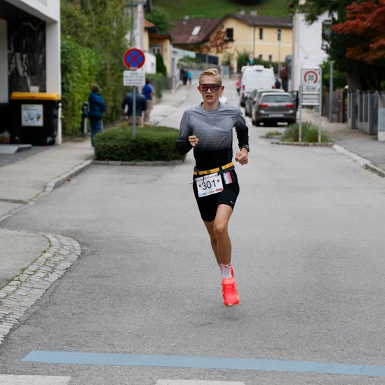 Ein Läufer mit einem Nummer 301 gekennzeichneten Startnummer läuft auf einer Straße. Auf beiden Seiten der Straße befinden sich Gebäude, Mülleimer und geparkte Autos.