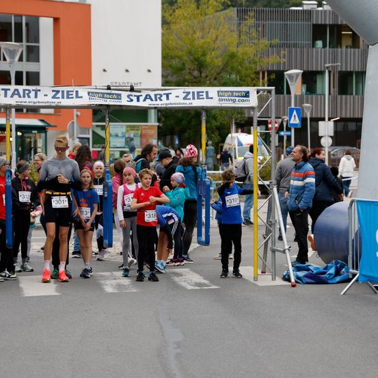 Teilnehmer eines Marathon-Events stehen an der Startlinie, tragen Nummern und Turnschuhe, mit einem Banner, das 'Start-Ziel' besagt. Die Umgebung ist im Freien mit Gebäuden und Bäumen im Hintergrund.