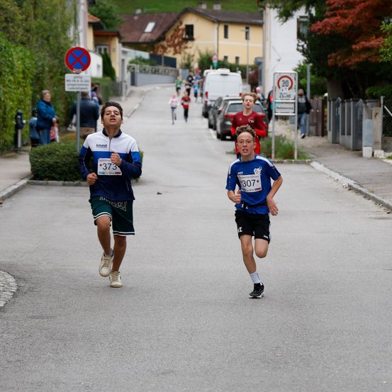 Zwei junge Läufer nehmen an einem Rennen auf einer Straße teil, mit Häusern, geparkten Autos und Bäumen im Hintergrund. Ein Läufer trägt ein blaues Trikot mit der Nummer 373, während der andere ein blaues Trikot mit der Nummer 307 trägt.