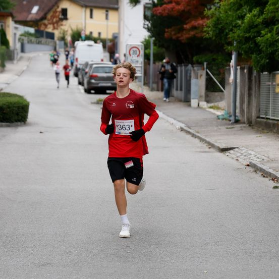 Ein Läufer in einem roten Trikot mit der Nummer 363 sprintet während eines Rennens eine Stadtstraße hinunter, mit anderen Läufern, geparkten Autos und Zuschauern im Hintergrund.