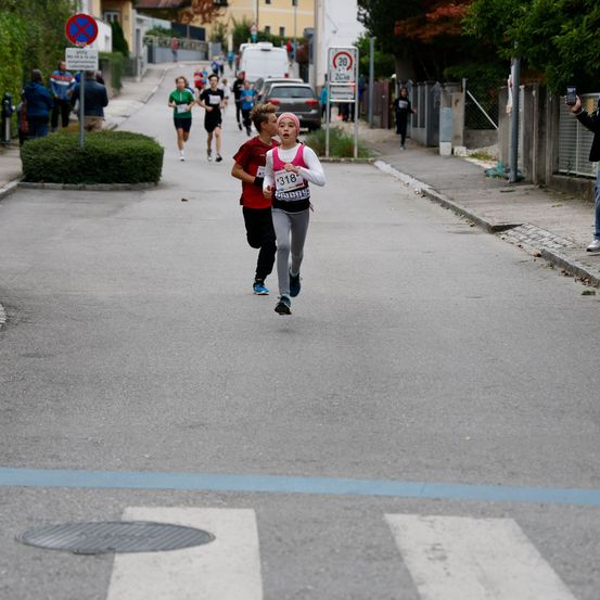 Eine Gruppe von Menschen nimmt an einem Laufrennen auf einer Straße mit einem Geschwindigkeitslimit von 30 km/h teil. Eine Frau mit der Nummer 318 auf ihrem Trikot führt das Rennen an.