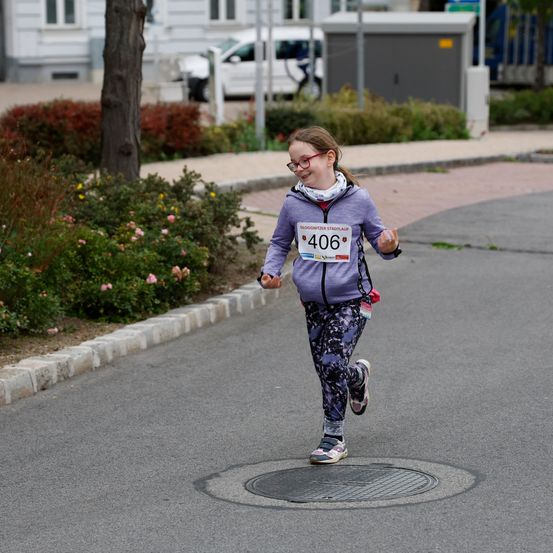 Ein junges Mädchen mit Brille und einem Nummernschild 406 läuft auf einer asphaltierten Straße, möglicherweise bei einem Rennen.