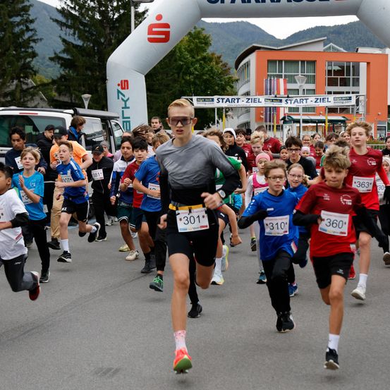 Marathonteilnehmer mit Nummern laufen in einem Rennen mit einem Start-Ziel-Banner. Sie sind von Zuschauern umgeben, mit Bäumen und einem Gebäude im Hintergrund.
