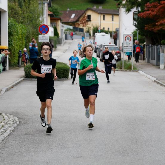 Mehrere Kinder laufen in einem Marathon auf einer Stadtstraße. Ein Junge mit Brille und der Nummer 333 und ein Mädchen mit der Nummer 379 führen das Rennen an.