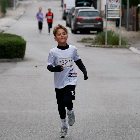 Ein junger Junge läuft in einem Rennen und trägt ein weißes Trikot mit der Nummer 321. Dahinter nehmen auch zwei andere Läufer am Rennen teil.